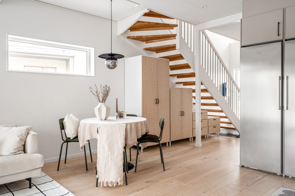 Ljus matplats efter homestyling med rundt bord, eleganta stolar, beige textilier och modern förvaring vid fönstret och trappa i radhus