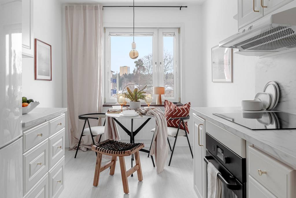 Ljust och modernt kök med vita skåp, rund matplats, mjuka textilier och stora fönster efter homestyling.