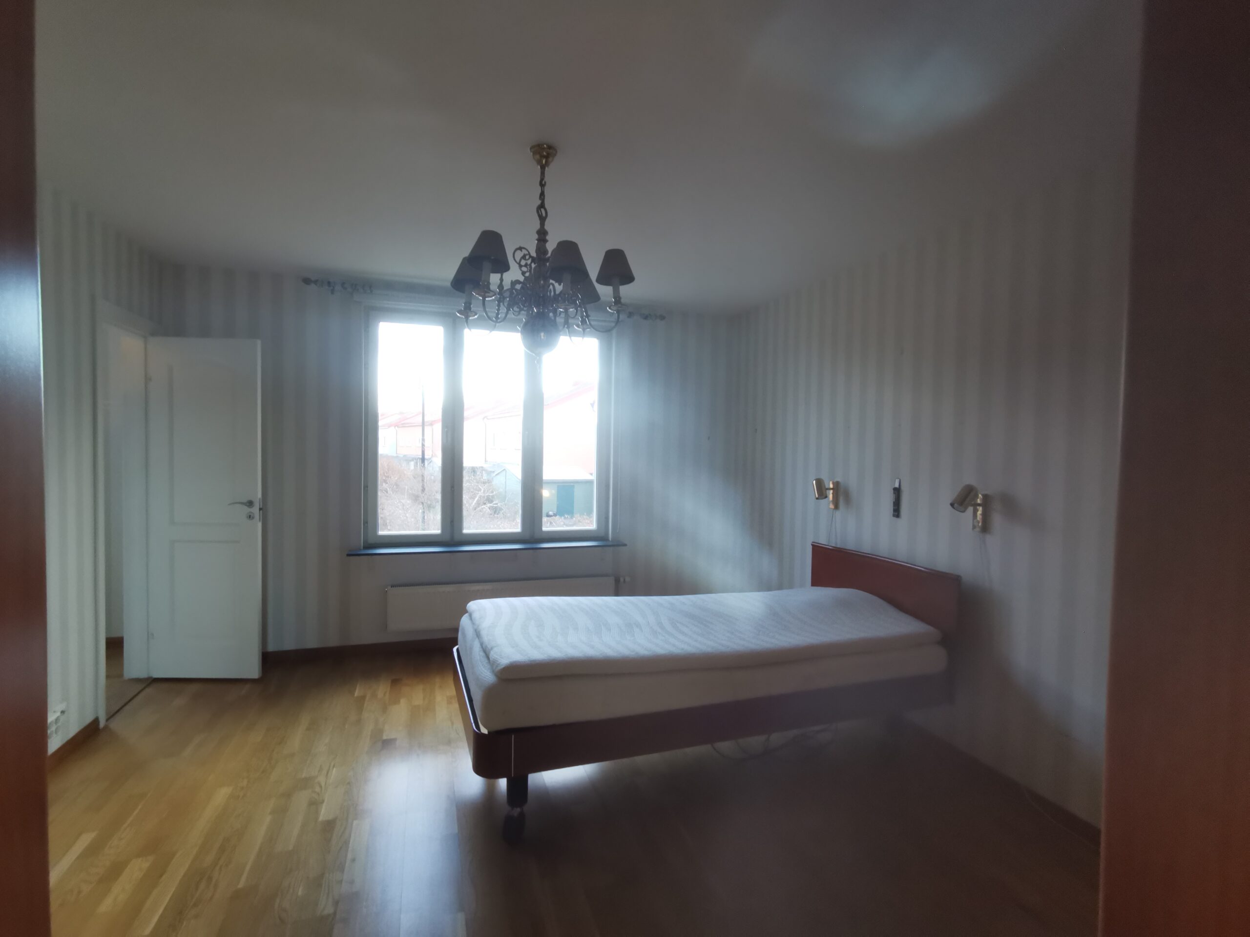 Sovrum före homestyling med enkel säng, tomma väggar, trägolv och stort fönster.