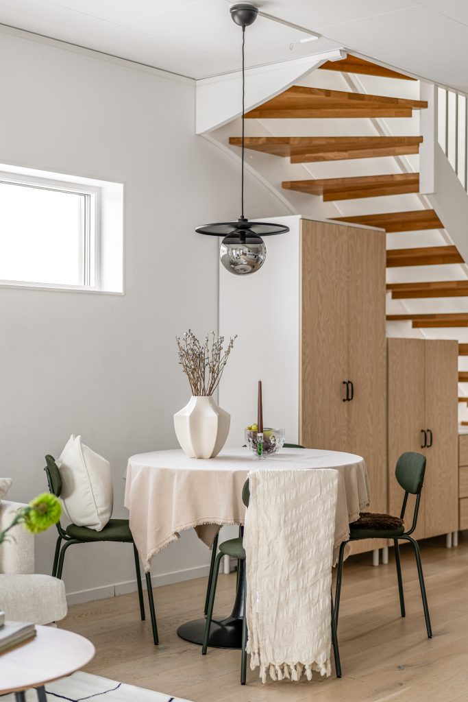 Ljus matplats efter homestyling med rundt bord, eleganta stolar, beige textilier och modern förvaring vid trappa i radhus.